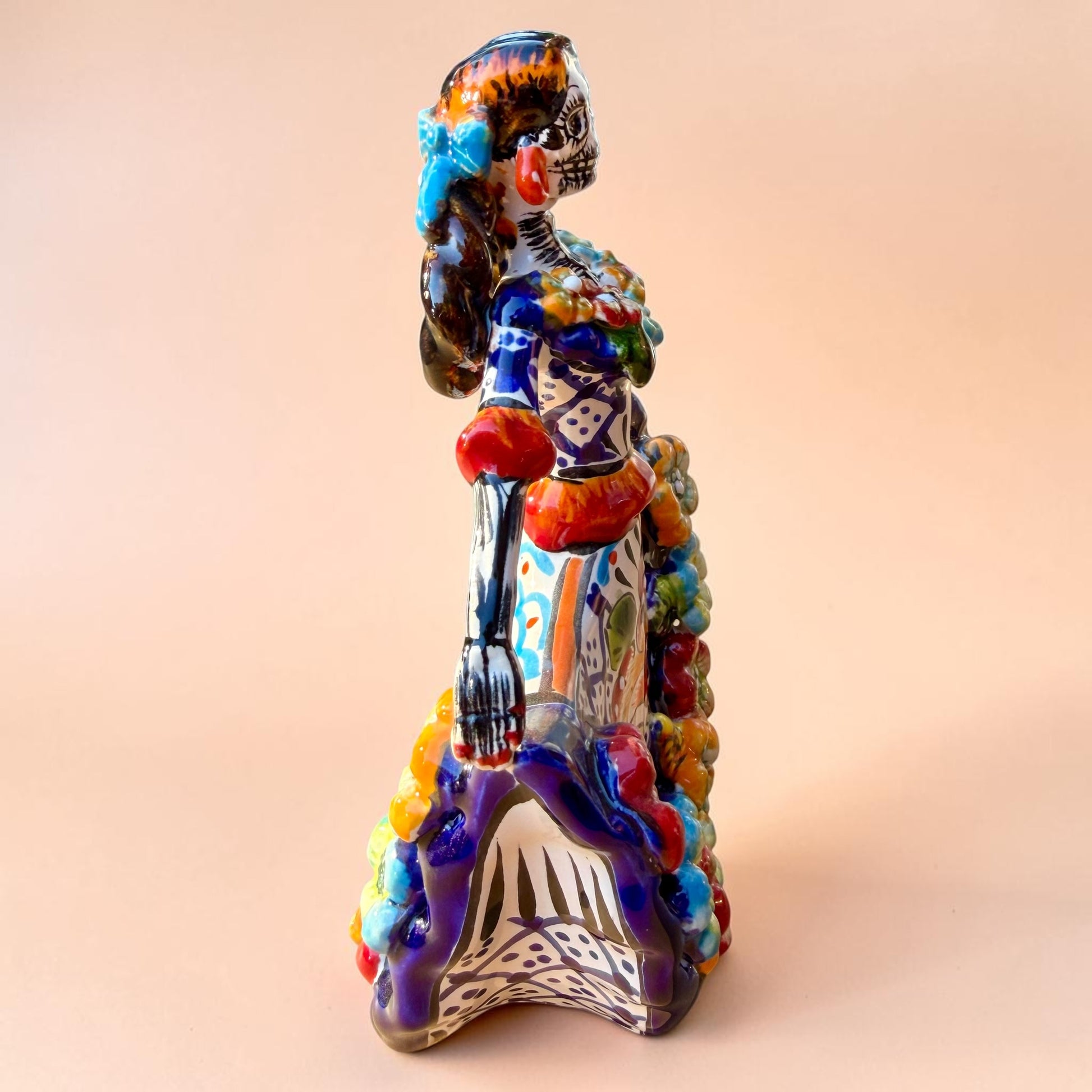 A colorful hand-painted Day of the Dead (Día de los Muertos) ceramic figurine of a  ballerina, decorated with vibrant floral patterns and a white skull head with braided hair, set against a solid peach background. Left side view of the doll showing her hand.
