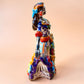 A colorful hand-painted Day of the Dead (Día de los Muertos) ceramic figurine of a  ballerina, decorated with vibrant floral patterns and a white skull head with braided hair, set against a solid peach background. Left side view of the doll showing her hand.