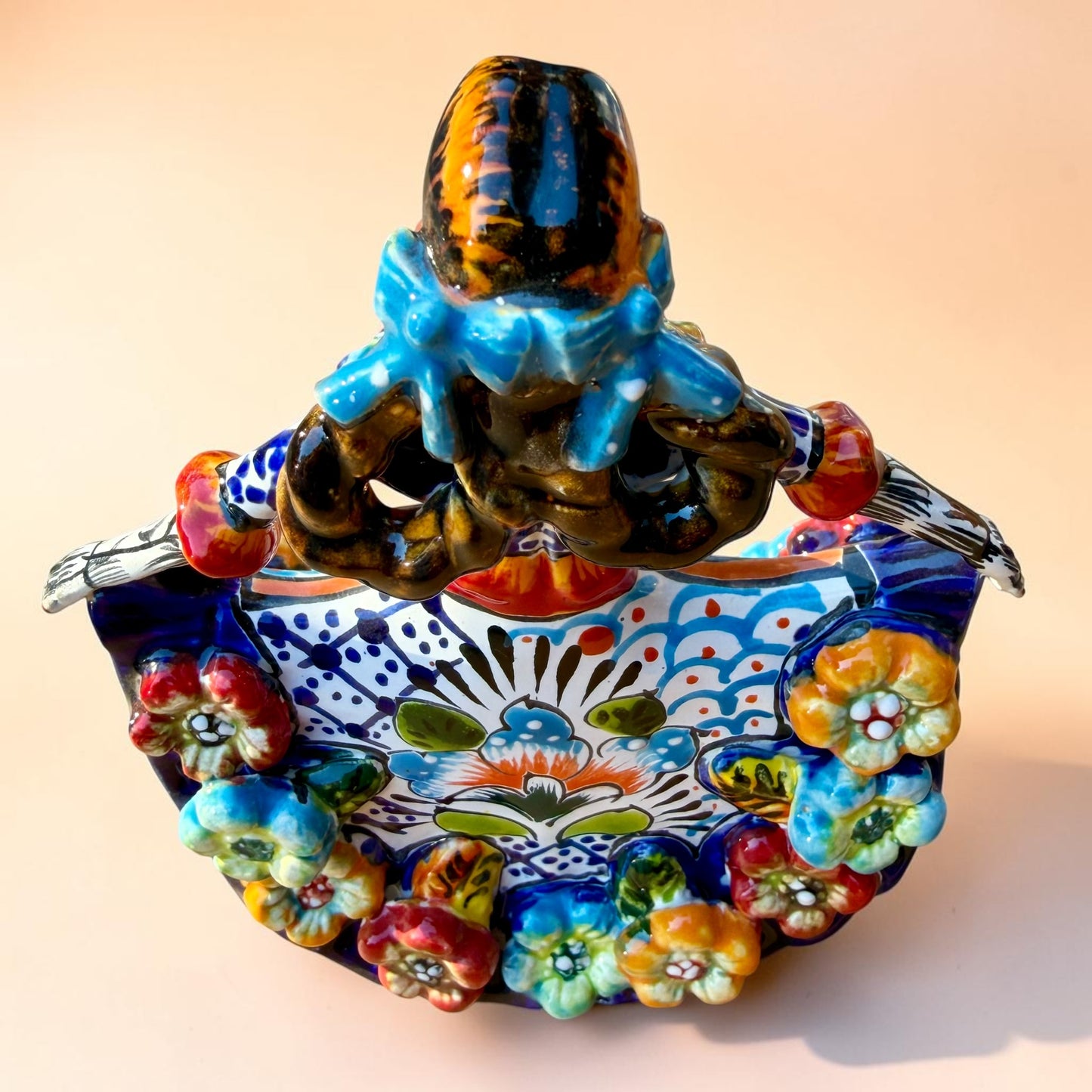 A colorful hand-painted Day of the Dead (Día de los Muertos) ceramic figurine of a  ballerina, decorated with vibrant floral patterns and a white skull head with braided hair, set against a solid peach background. Top back view showing her hair bows.