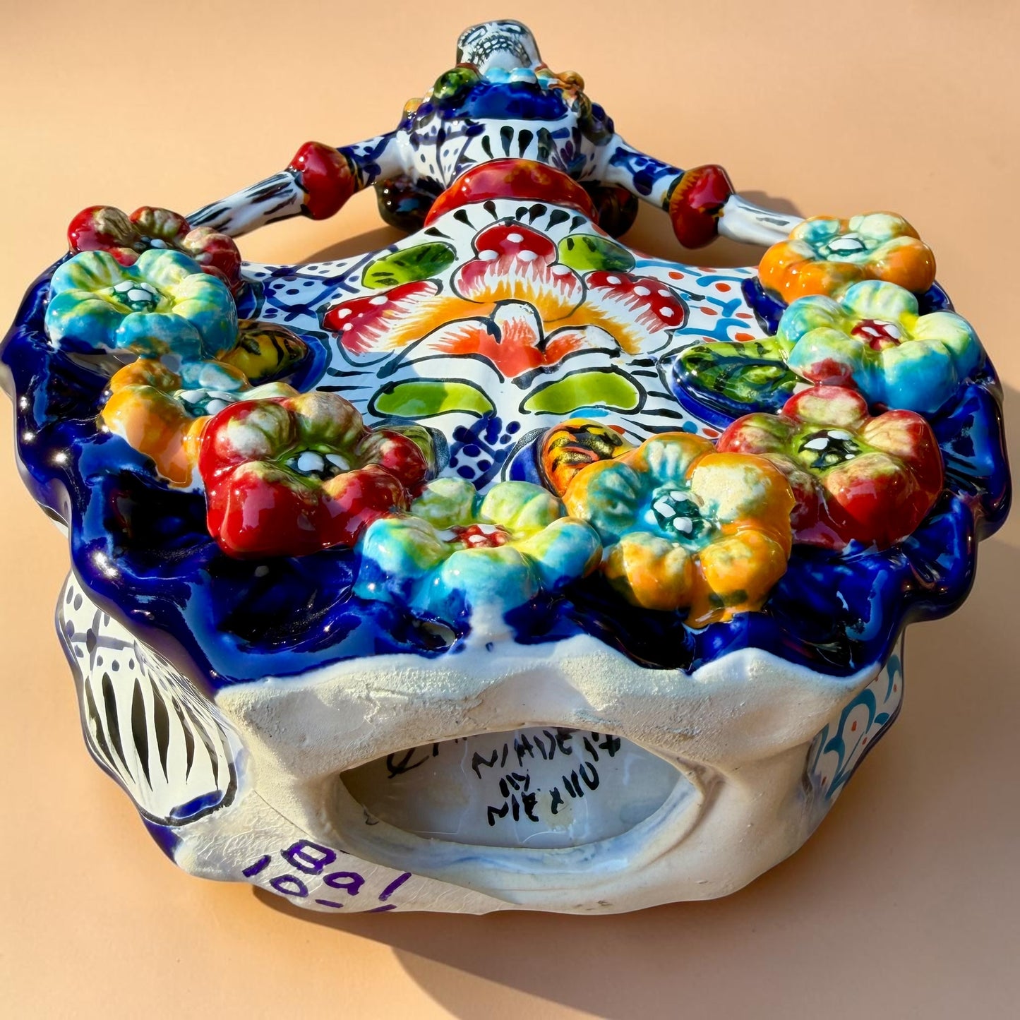 A colorful hand-painted Day of the Dead (Día de los Muertos) ceramic figurine of a  ballerina, decorated with vibrant floral patterns and a white skull head with braided hair, set against a solid peach background. Bottom of figurine showing artist signature.