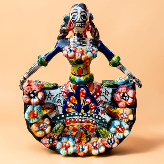 A colorful hand-painted Day of the Dead (Día de los Muertos) ceramic figurine of a  ballerina, decorated with vibrant floral patterns and a white skull head with braided hair, set against a solid peach background.