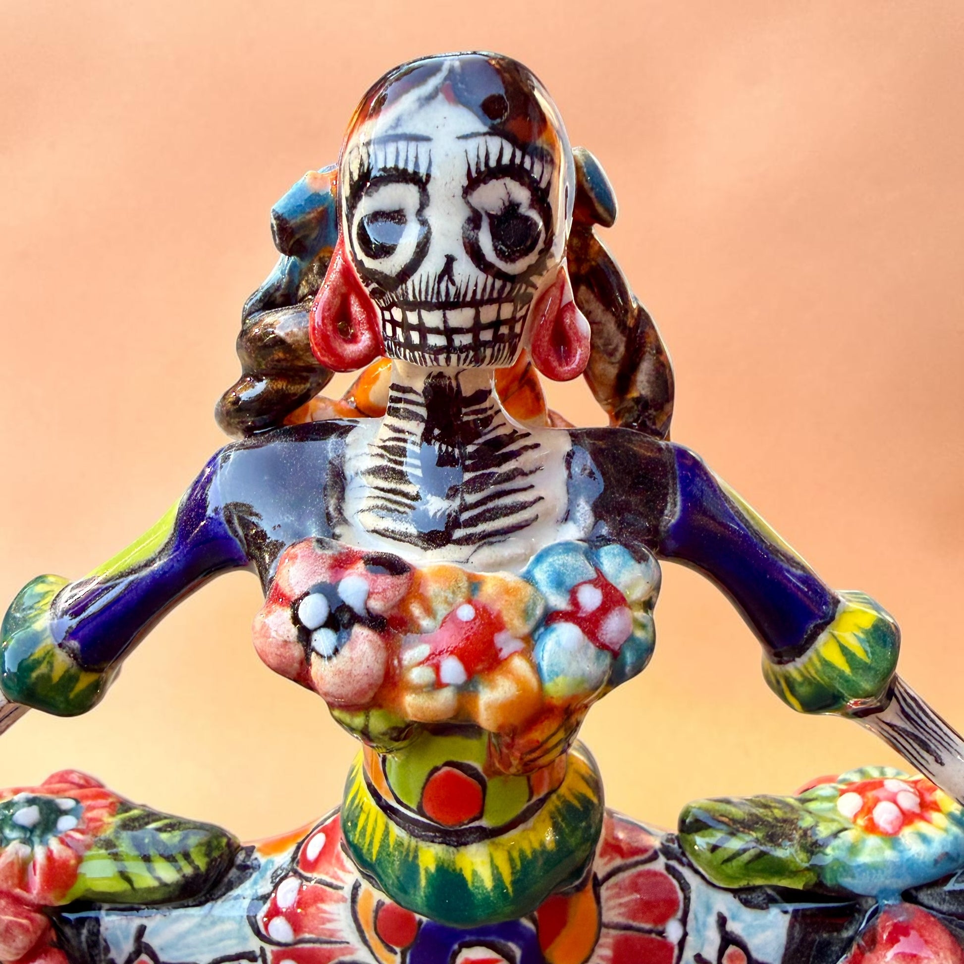 A colorful hand-painted Day of the Dead (Día de los Muertos) ceramic figurine of a  ballerina, decorated with vibrant floral patterns and a white skull head with braided hair, set against a solid peach background. Close up of face.