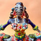 A colorful hand-painted Day of the Dead (Día de los Muertos) ceramic figurine of a  ballerina, decorated with vibrant floral patterns and a white skull head with braided hair, set against a solid peach background. Close up of face.