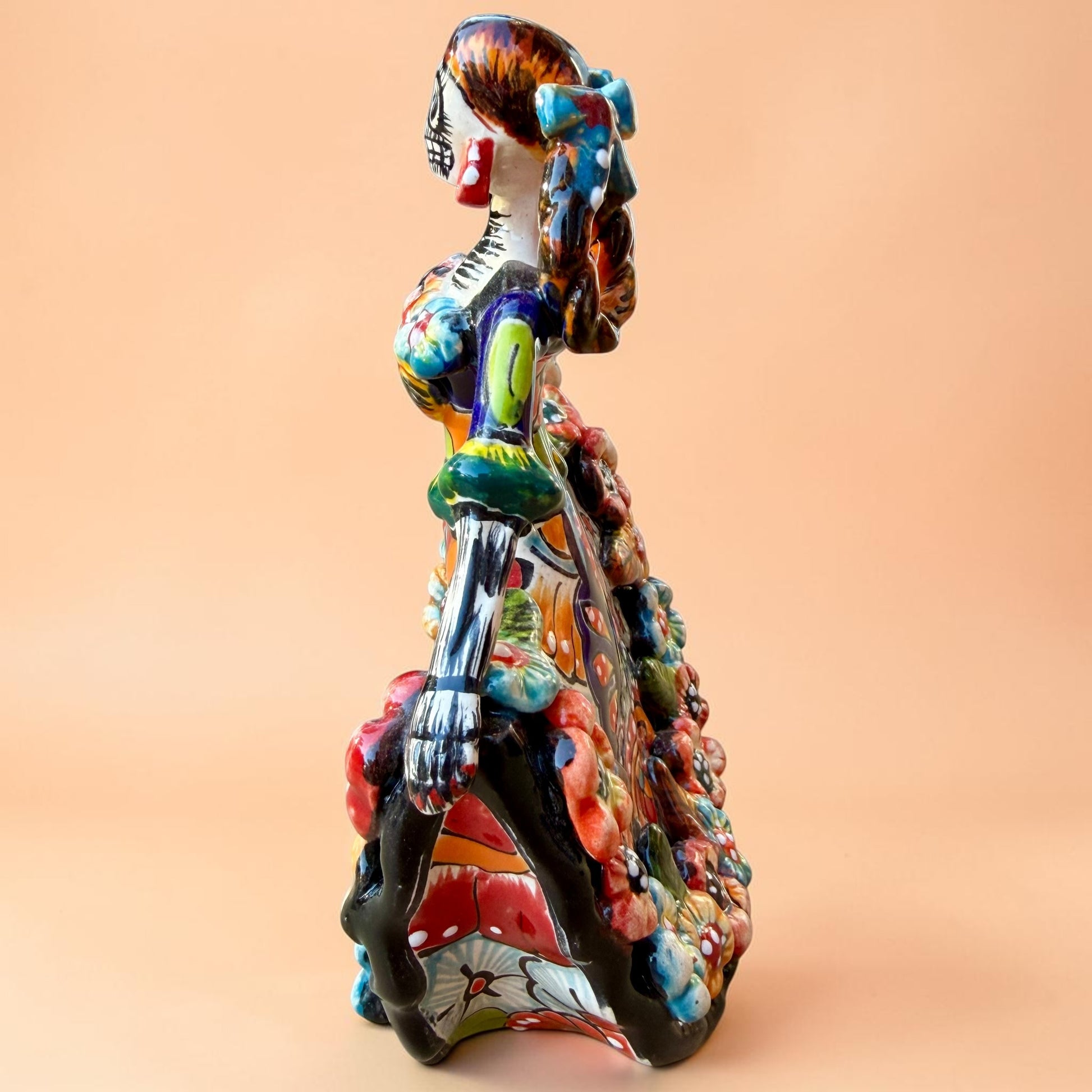 A colorful hand-painted Day of the Dead (Día de los Muertos) ceramic figurine of a  ballerina, decorated with vibrant floral patterns and a white skull head with braided hair, set against a solid peach background. Right side view of ballerina.