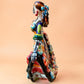 A colorful hand-painted Day of the Dead (Día de los Muertos) ceramic figurine of a  ballerina, decorated with vibrant floral patterns and a white skull head with braided hair, set against a solid peach background. Right side view of ballerina.