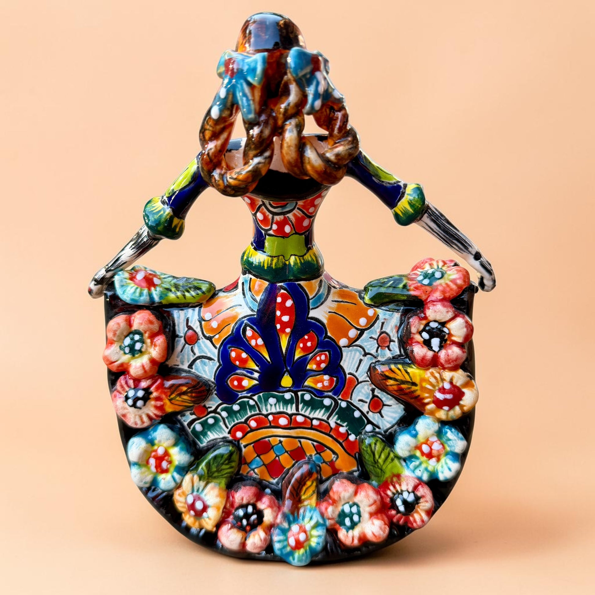 A colorful hand-painted Day of the Dead (Día de los Muertos) ceramic figurine of a  ballerina, decorated with vibrant floral patterns and a white skull head with braided hair, set against a solid peach background. Back side view of Ballerina doll.