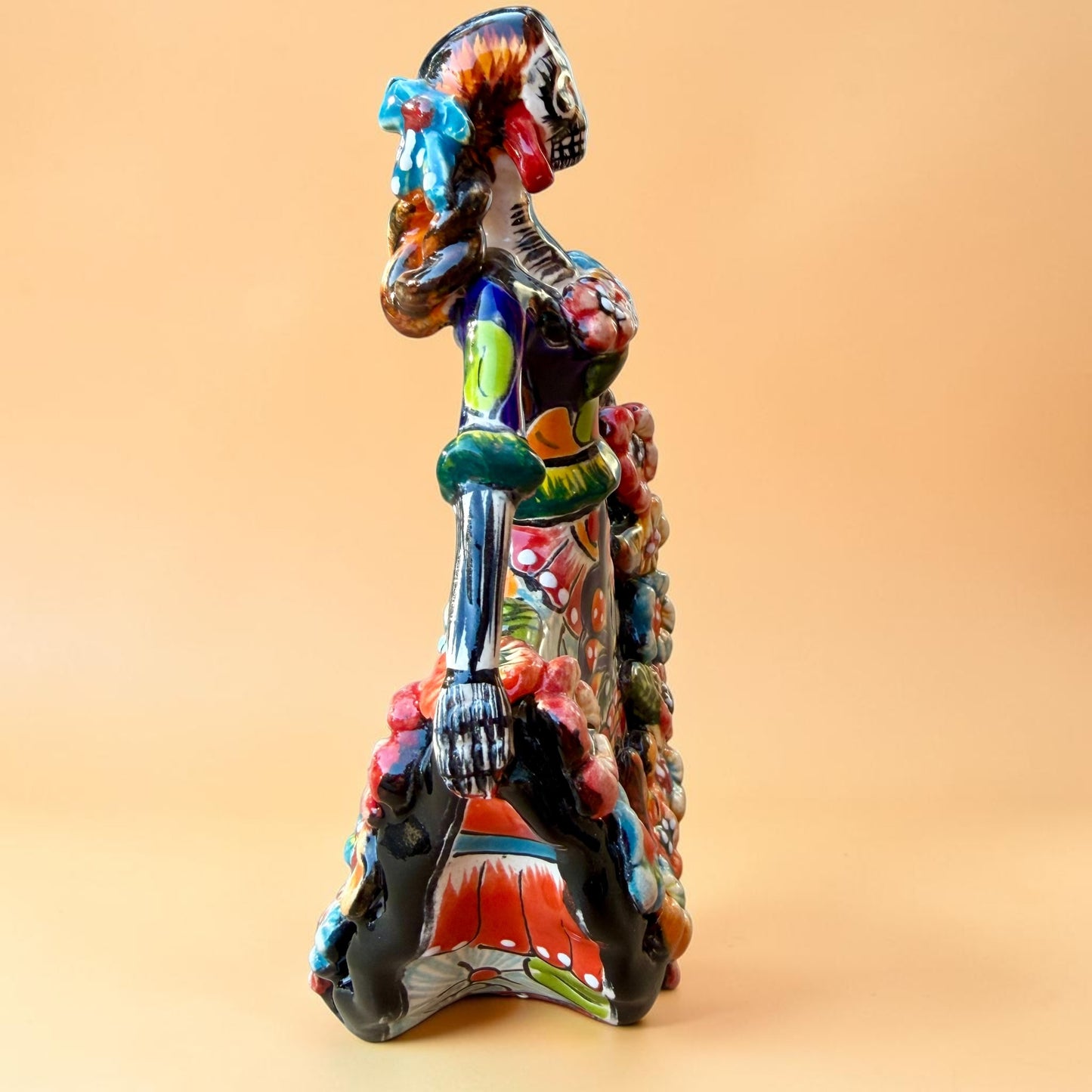 A colorful hand-painted Day of the Dead (Día de los Muertos) ceramic figurine of a  ballerina, decorated with vibrant floral patterns and a white skull head with braided hair, set against a solid peach background. Left side view of ballerina catrina doll.