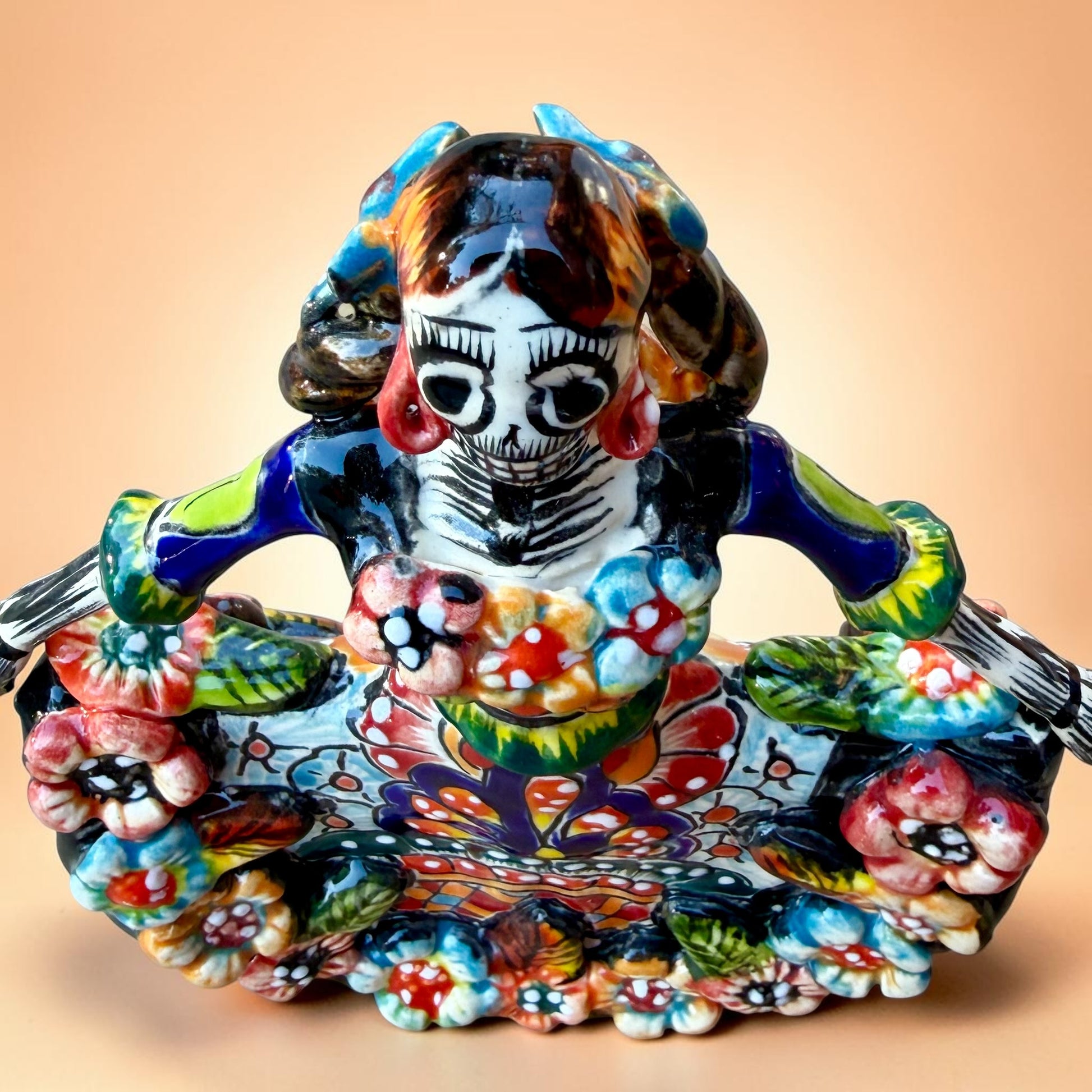 A colorful hand-painted Day of the Dead (Día de los Muertos) ceramic figurine of a  ballerina, decorated with vibrant floral patterns and a white skull head with braided hair, set against a solid peach background. Top view of doll looking down.