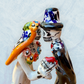 A hand-painted ceramic Day of the Dead figurine of a dancing skeleton couple, featuring a bride and groom with vibrant multicolor floral patterns on a white background. Close of of the couples faces.