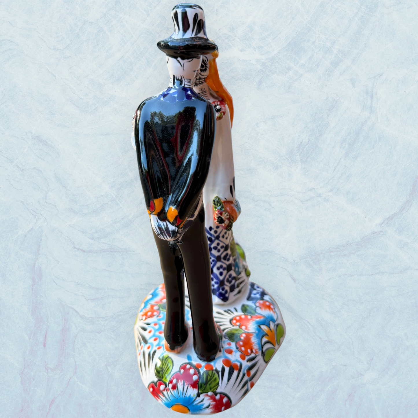 A hand-painted ceramic Day of the Dead figurine of a dancing skeleton couple, featuring a bride and groom with vibrant multicolor floral patterns on a white background.  side view focusing on the groom.