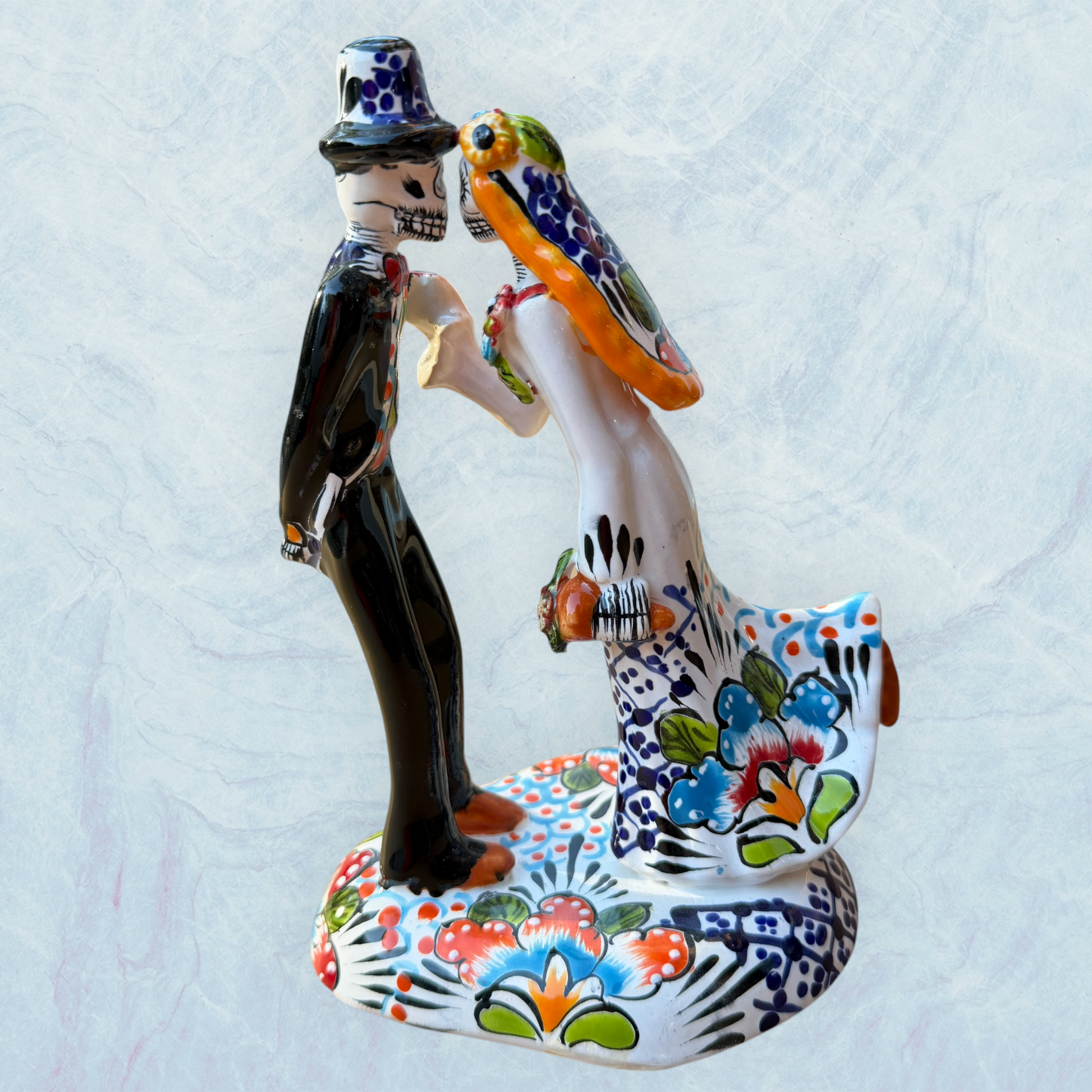 A hand-painted ceramic Day of the Dead figurine of a dancing skeleton couple, featuring a bride and groom with vibrant multicolor floral patterns on a white background. View of the opposite side of the figurine.