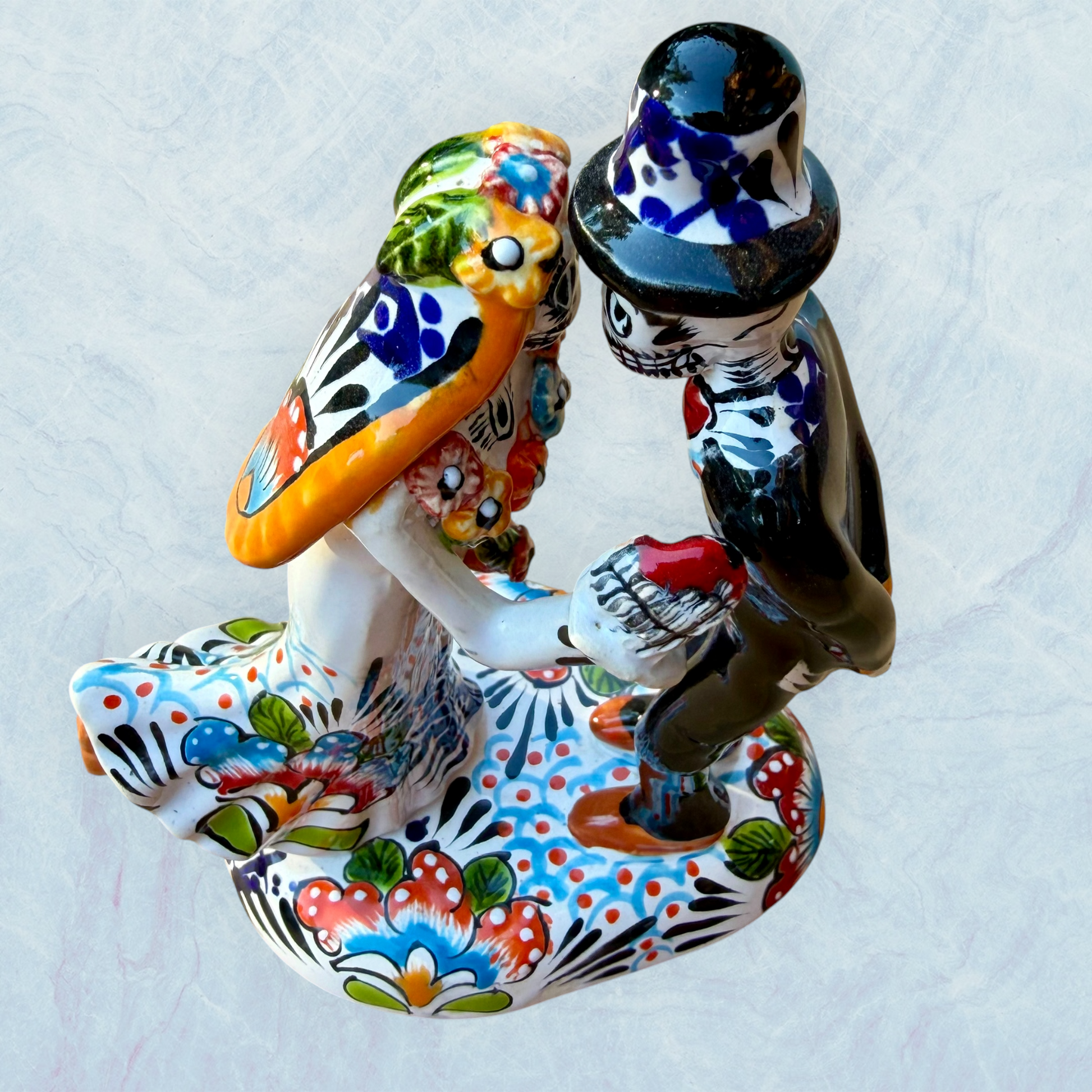 A hand-painted ceramic Day of the Dead figurine of a dancing skeleton couple, featuring a bride and groom with vibrant multicolor floral patterns on a white background. Top view.