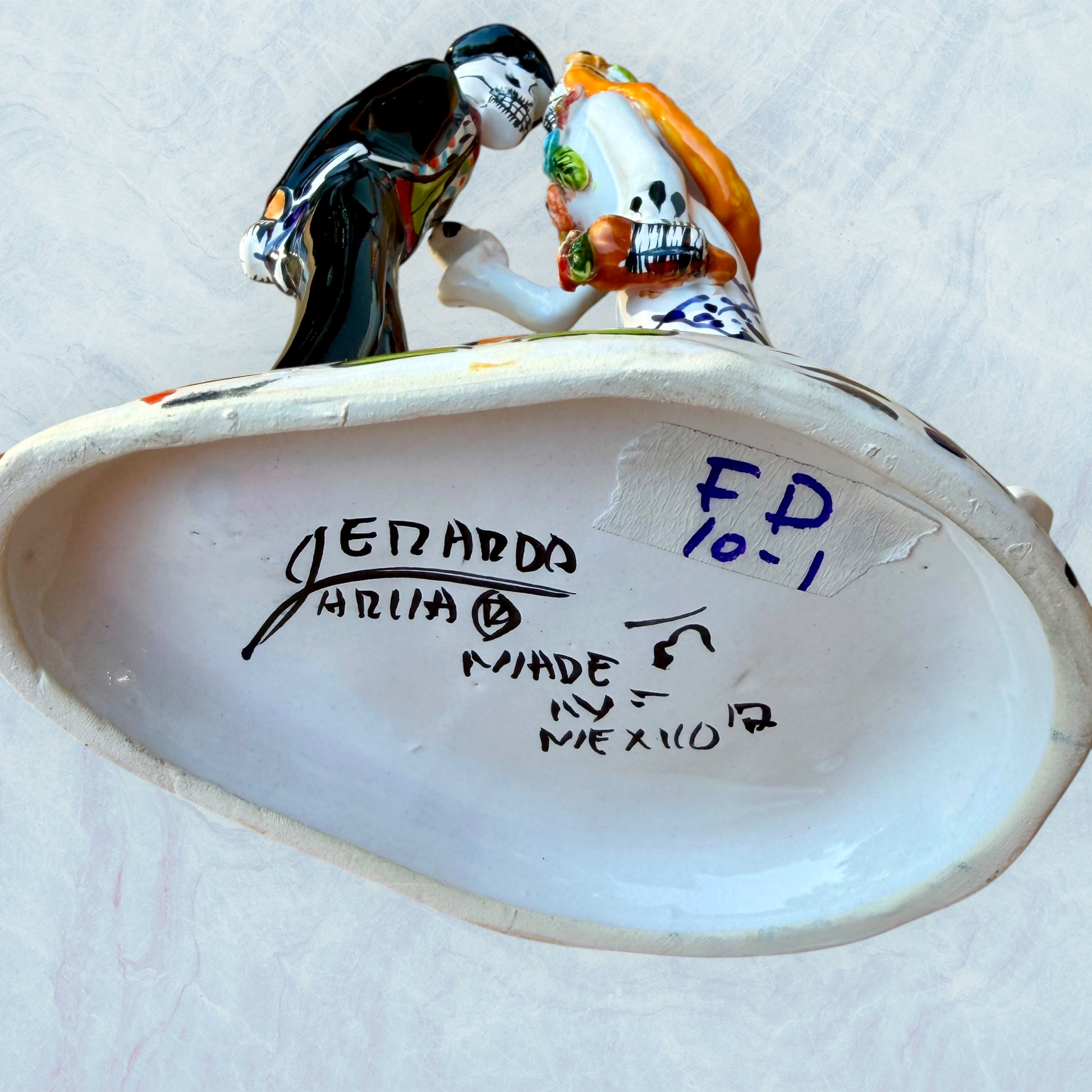 A hand-painted ceramic Day of the Dead figurine of a dancing skeleton couple, featuring a bride and groom with vibrant multicolor floral patterns on a white background. Bottom view showing artist signature.