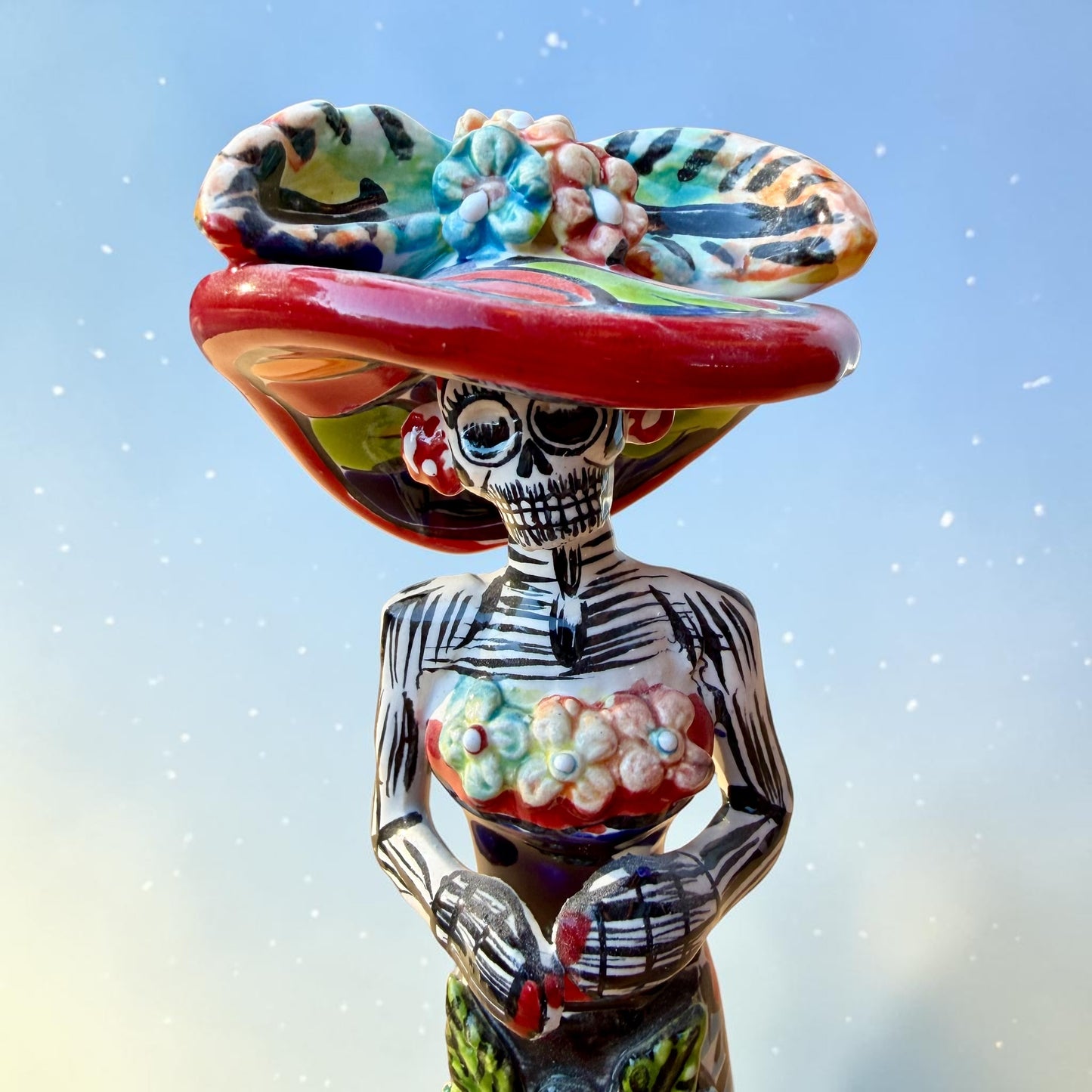 Mexican Catrina Doll made out of Talavera with Calla Lillies on the front of her dress.  Colorful pieces and a big feathered hat and a big toothy smile. Close up of figurines face.