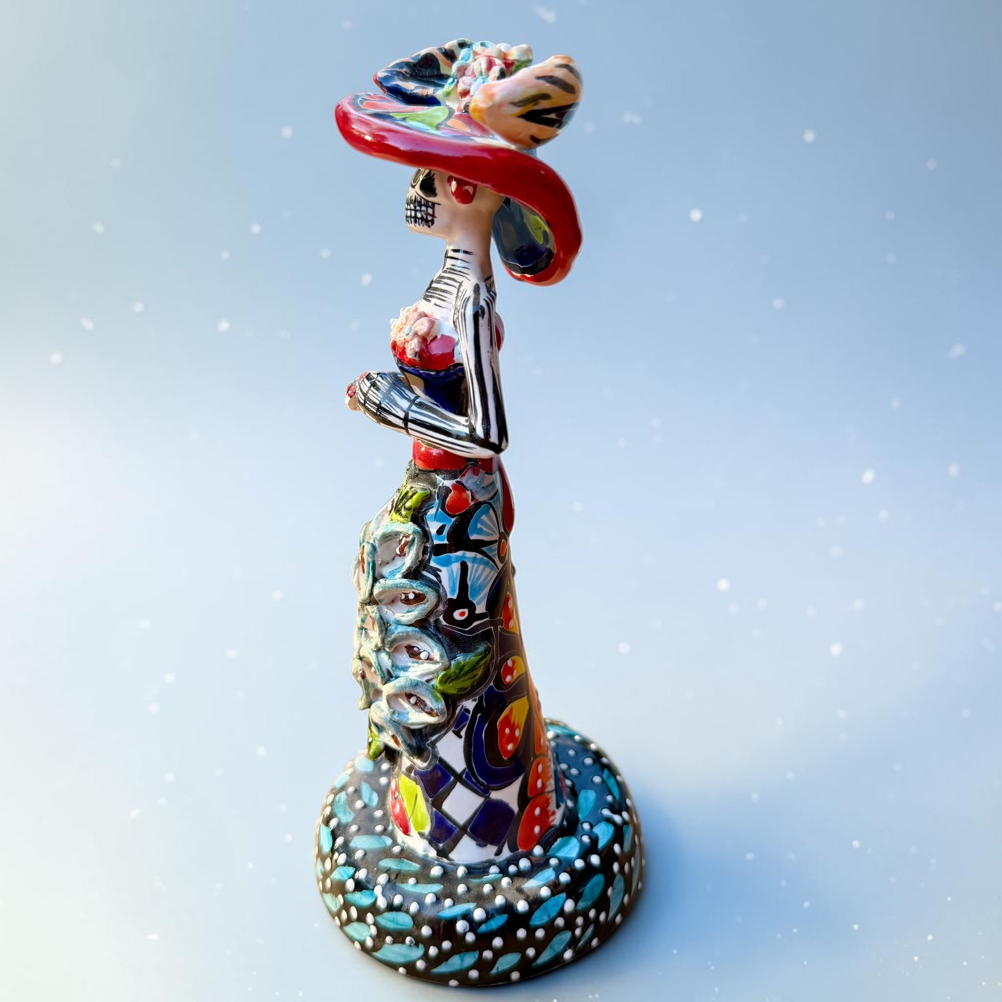 Mexican Catrina Doll made out of Talavera with Calla Lillies on the front of her dress.  Colorful pieces and a big feathered hat and a big toothy smile. Right side view of the catrina doll.