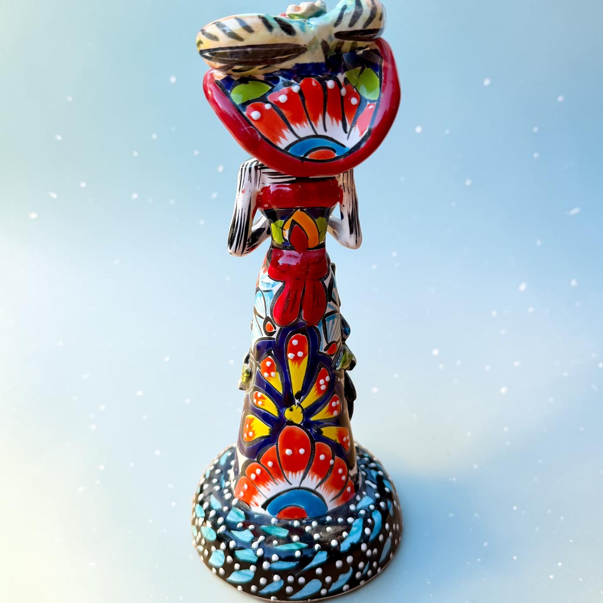 Mexican Catrina Doll made out of Talavera with Calla Lillies on the front of her dress.  Colorful pieces and a big feathered hat and a big toothy smile. Back view of catrina doll with big bow around her waist.