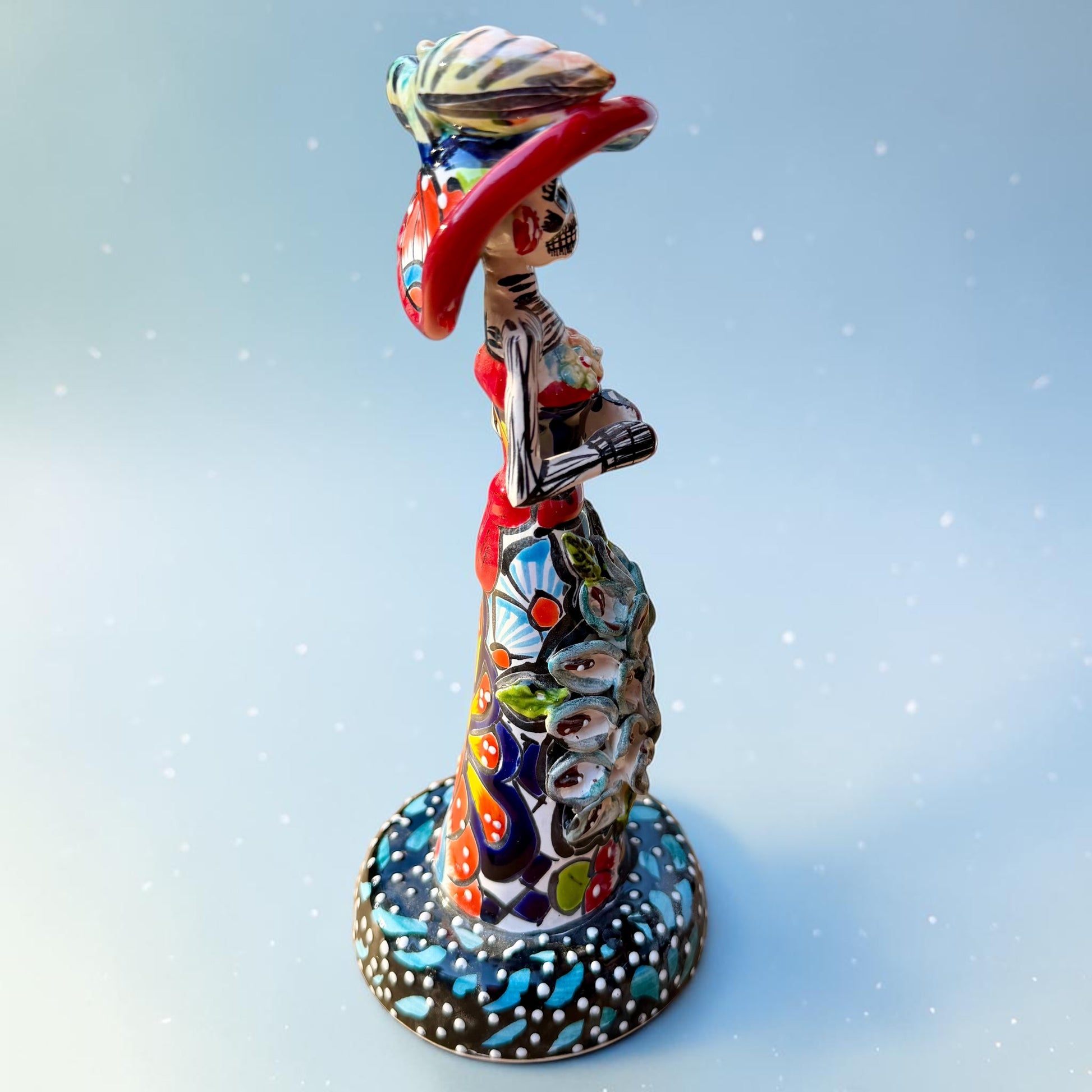 Mexican Catrina Doll made out of Talavera with Calla Lillies on the front of her dress.  Colorful pieces and a big feathered hat and a big toothy smile. left side view of catrina.