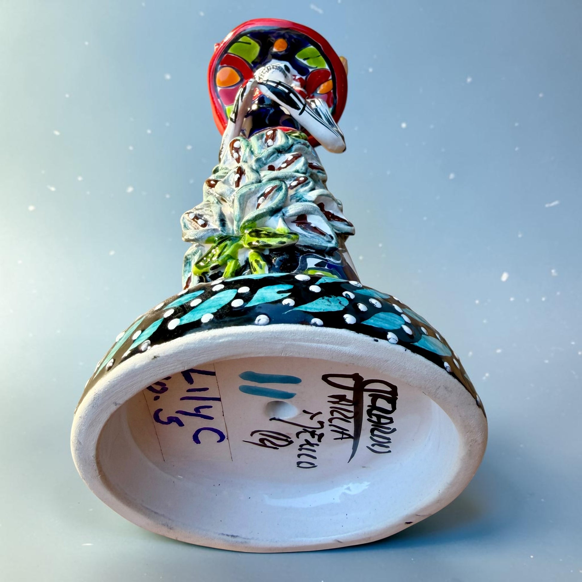 Mexican Catrina Doll made out of Talavera with Calla Lillies on the front of her dress.  Colorful pieces and a big feathered hat and a big toothy smile. Bottom view showing signature on the base.