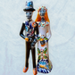 A decorative ceramic Day of the Dead wedding couple figurine featuring a black-and-white skeleton groom in a suit and hat with a butterfly patterned hat, and a white bride with orange hair in a floral gown with butterflies holding a bouquet, set against a light textured background.