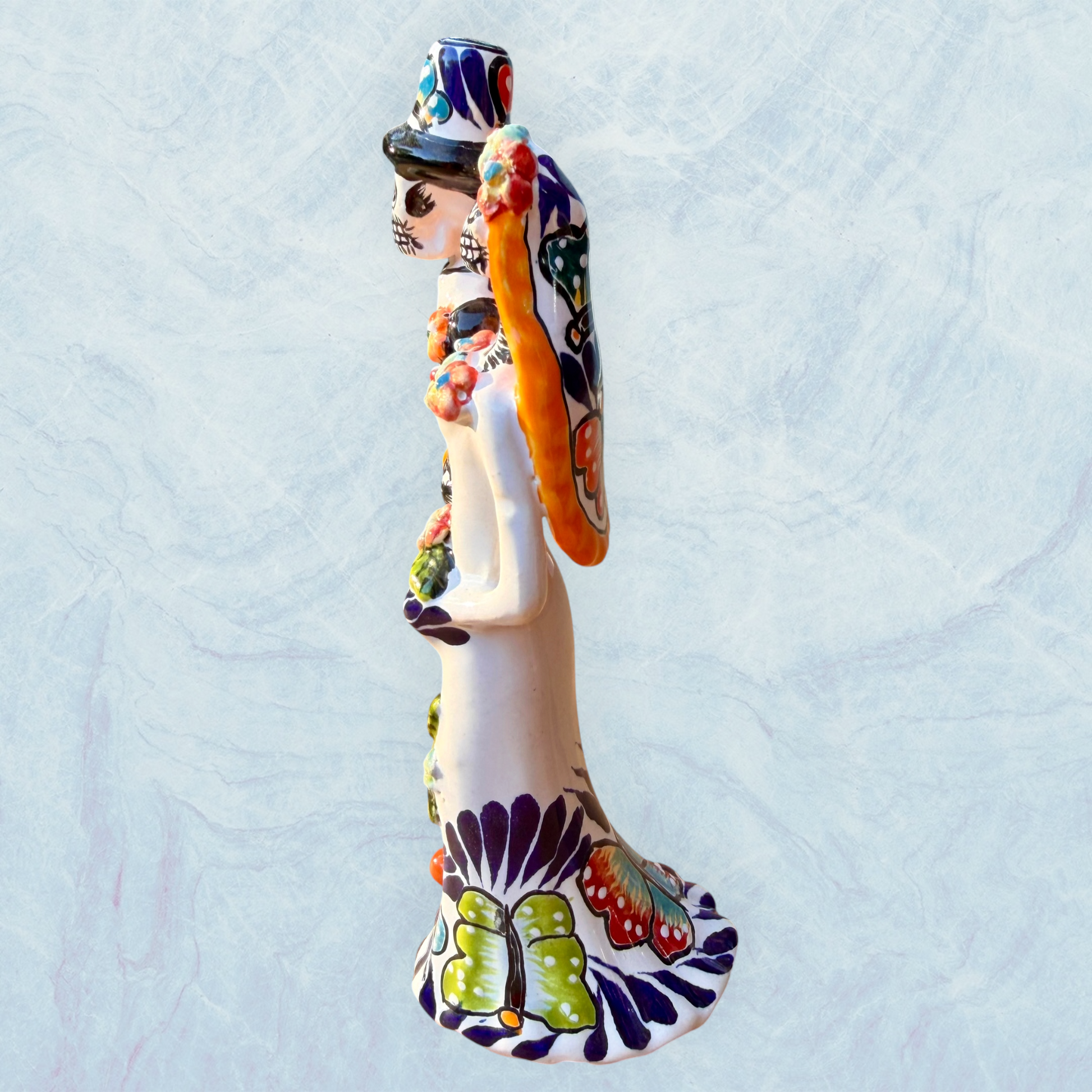 A decorative ceramic Day of the Dead wedding couple figurine featuring a black-and-white skeleton groom in a suit and hat with a butterfly patterned hat, and a white bride with orange hair in a floral gown with butterflies holding a bouquet, set against a light textured background. This is the right side view.
