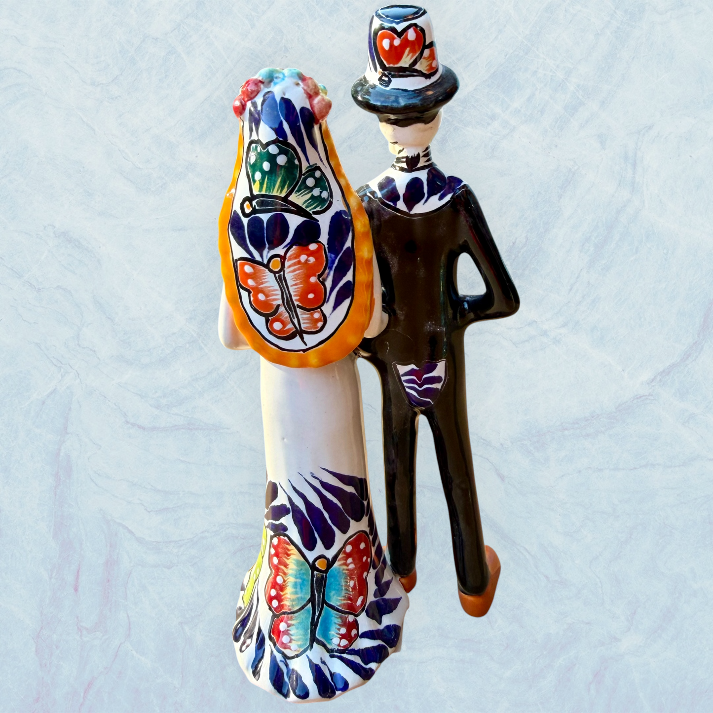 A decorative ceramic Day of the Dead wedding couple figurine featuring a black-and-white skeleton groom in a suit and hat with a butterfly patterned hat, and a white bride with orange hair in a floral gown with butterflies holding a bouquet, set against a light textured background. This is the back view showing veil and butterfly decorations.