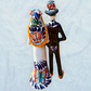 A decorative ceramic Day of the Dead wedding couple figurine featuring a black-and-white skeleton groom in a suit and hat with a butterfly patterned hat, and a white bride with orange hair in a floral gown with butterflies holding a bouquet, set against a light textured background. This is the back view showing veil and butterfly decorations.