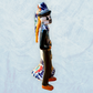 A decorative ceramic Day of the Dead wedding couple figurine featuring a black-and-white skeleton groom in a suit and hat with a butterfly patterned hat, and a white bride with orange hair in a floral gown with butterflies holding a bouquet, set against a light textured background. Left side view focusing on groom.