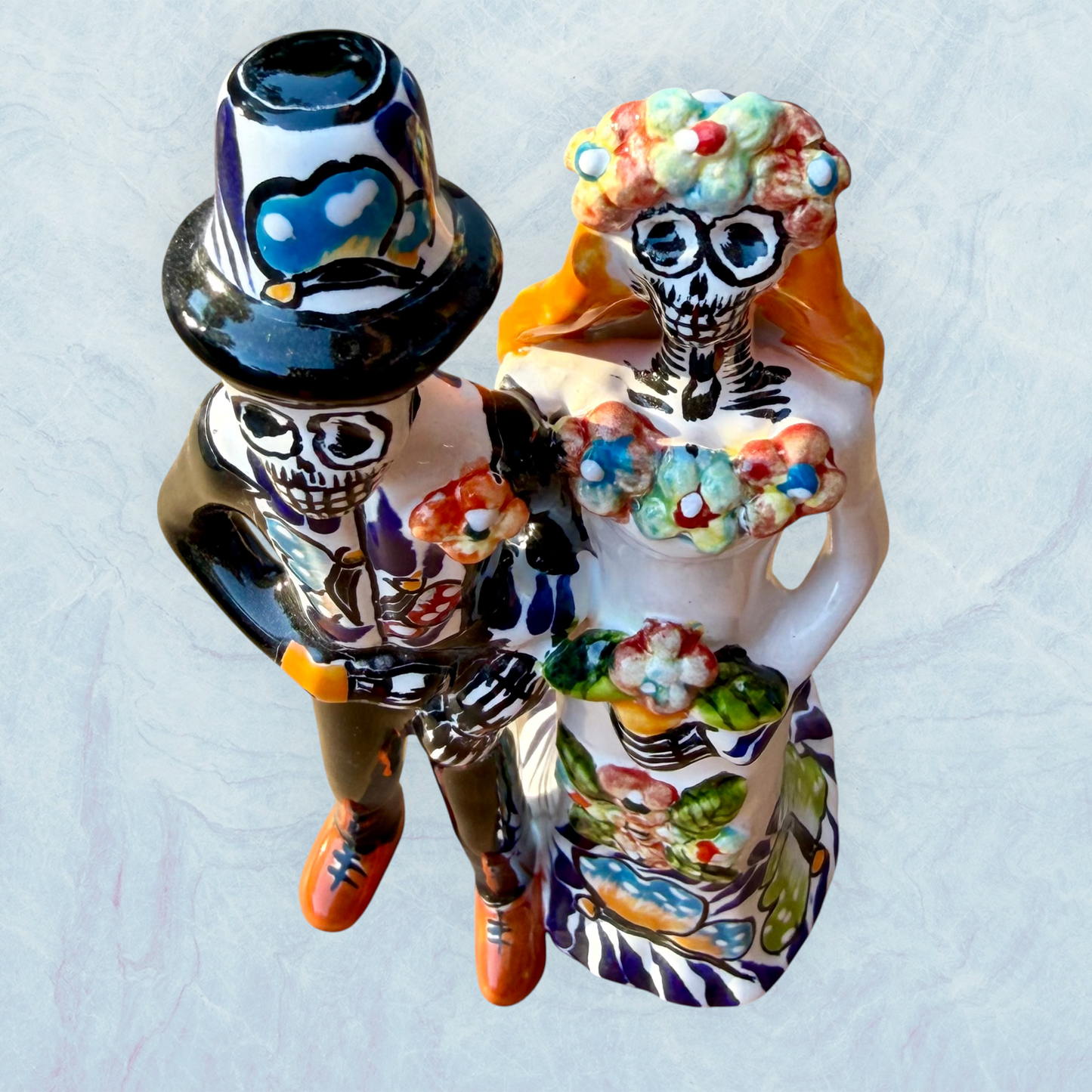 A decorative ceramic Day of the Dead wedding couple figurine featuring a black-and-white skeleton groom in a suit and hat with a butterfly patterned hat, and a white bride with orange hair in a floral gown with butterflies holding a bouquet, set against a light textured background. Top View.