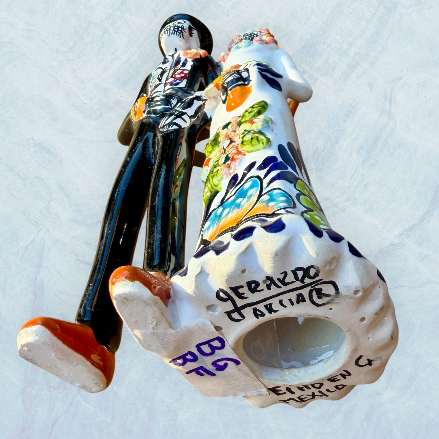 A decorative ceramic Day of the Dead wedding couple figurine featuring a black-and-white skeleton groom in a suit and hat with a butterfly patterned hat, and a white bride with orange hair in a floral gown with butterflies holding a bouquet, set against a light textured background. Bottom view showing artist signature.