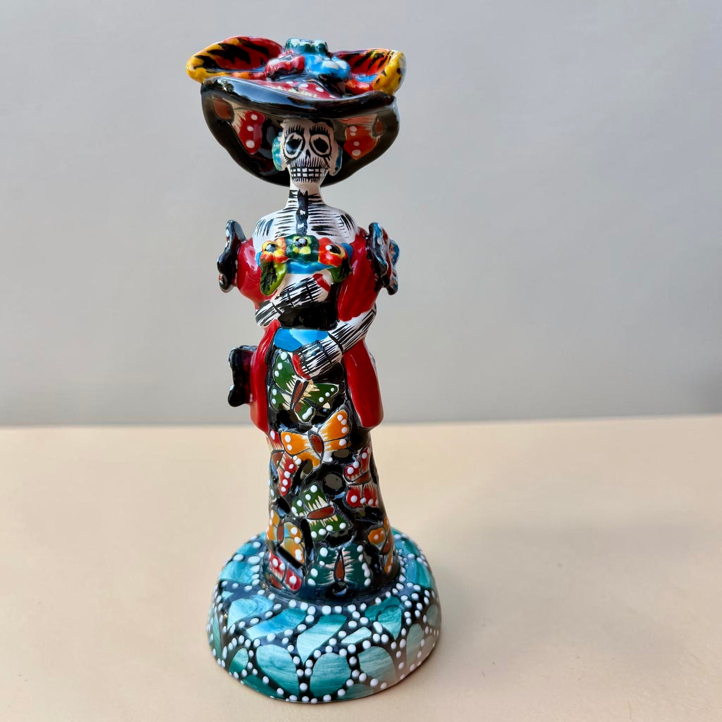 A colorful Day of the Dead (Día de los Muertos) skeleton figurine standing on a patterned base, decorated with  butterflies in vibrant red, blue, green, yellow, and black painted details.