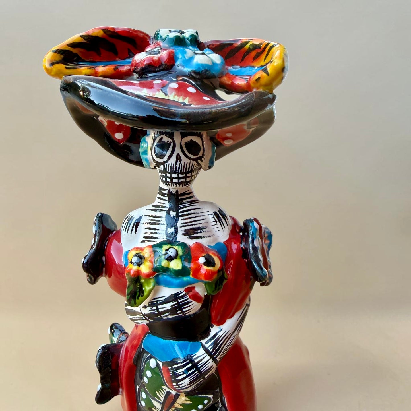 A colorful Day of the Dead (Día de los Muertos) skeleton figurine standing on a patterned base, decorated with  butterflies in vibrant red, blue, green, yellow, and black painted details. Close up of face with big hat.
