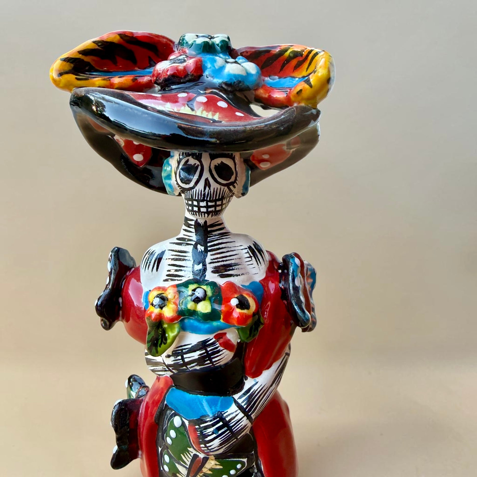 A colorful Day of the Dead (Día de los Muertos) skeleton figurine standing on a patterned base, decorated with  butterflies in vibrant red, blue, green, yellow, and black painted details. Close up of face with big hat.
