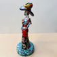 A colorful Day of the Dead (Día de los Muertos) skeleton figurine standing on a patterned base, decorated with  butterflies in vibrant red, blue, green, yellow, and black painted details. Right side views of doll.
