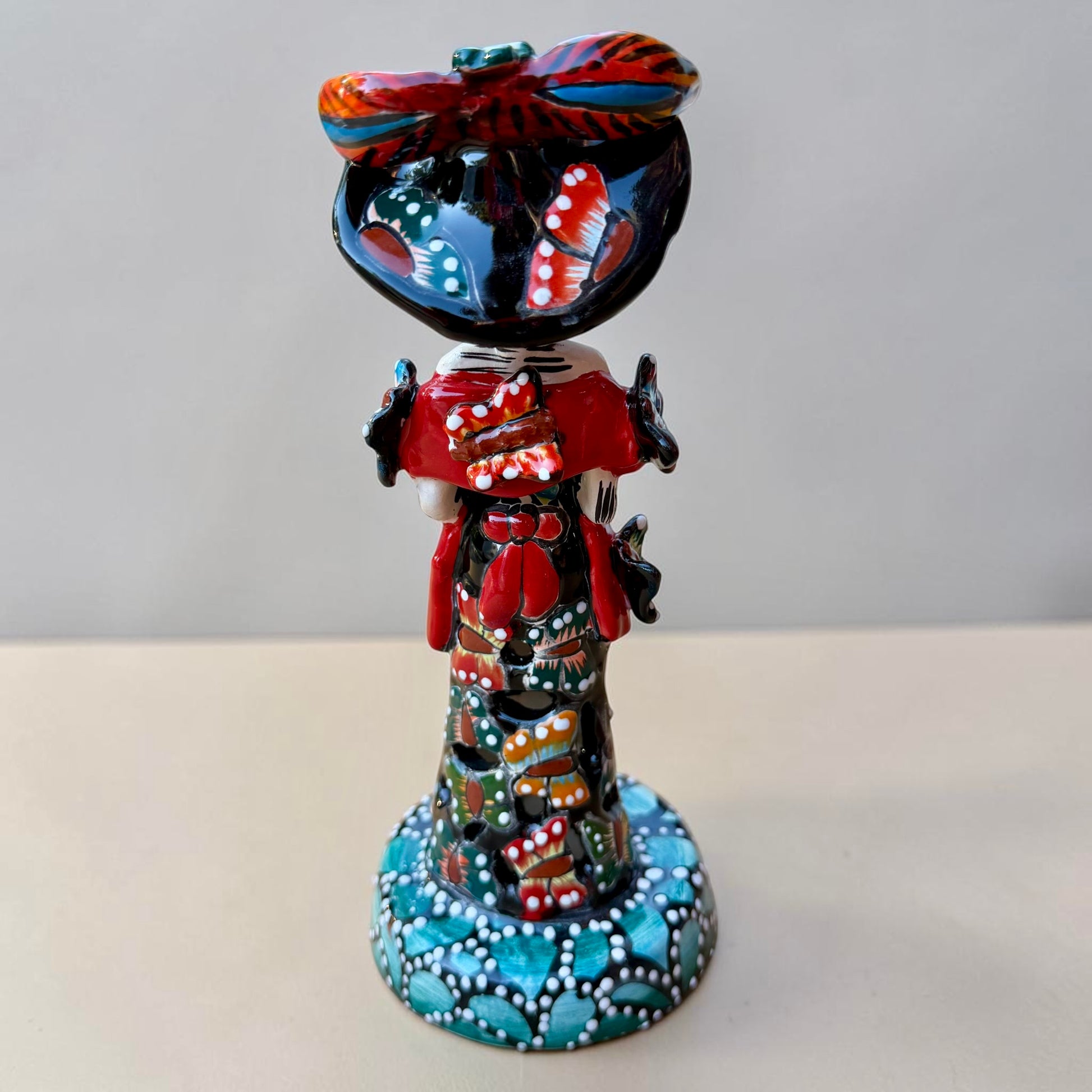 A colorful Day of the Dead (Día de los Muertos) skeleton figurine standing on a patterned base, decorated with  butterflies in vibrant red, blue, green, yellow, and black painted details. Back view of doll showing bow around her waist.