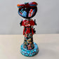 A colorful Day of the Dead (Día de los Muertos) skeleton figurine standing on a patterned base, decorated with  butterflies in vibrant red, blue, green, yellow, and black painted details. Back view of doll showing bow around her waist.