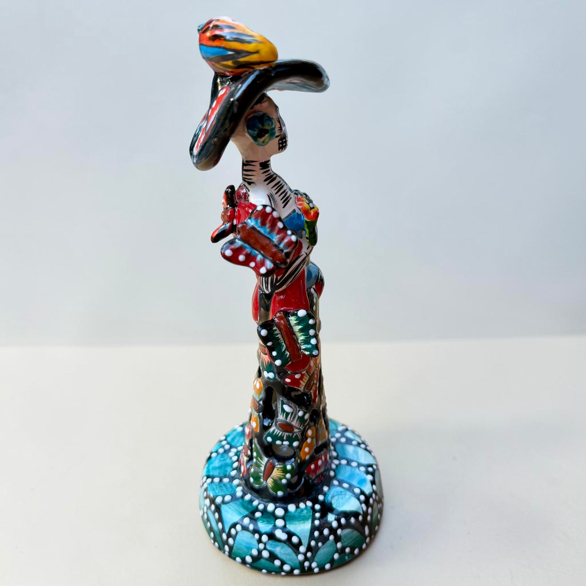 A colorful Day of the Dead (Día de los Muertos) skeleton figurine standing on a patterned base, decorated with  butterflies in vibrant red, blue, green, yellow, and black painted details. Left Side View of Doll
