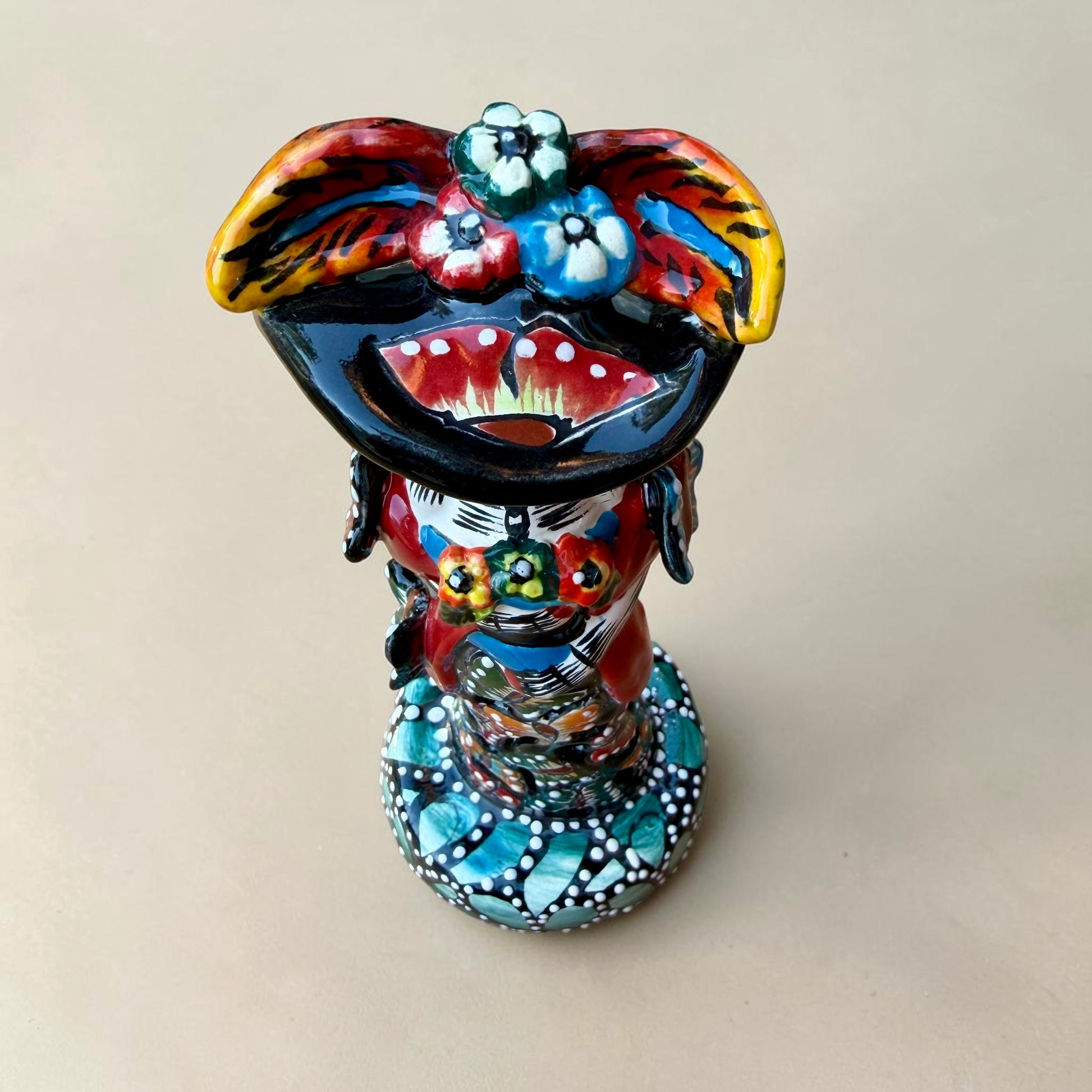 A colorful Day of the Dead (Día de los Muertos) skeleton figurine standing on a patterned base, decorated with  butterflies in vibrant red, blue, green, yellow, and black painted details. Top view of the figurine.