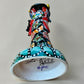 A colorful Day of the Dead (Día de los Muertos) skeleton figurine standing on a patterned base, decorated with  butterflies in vibrant red, blue, green, yellow, and black painted details. Bottom view showing artist signature.